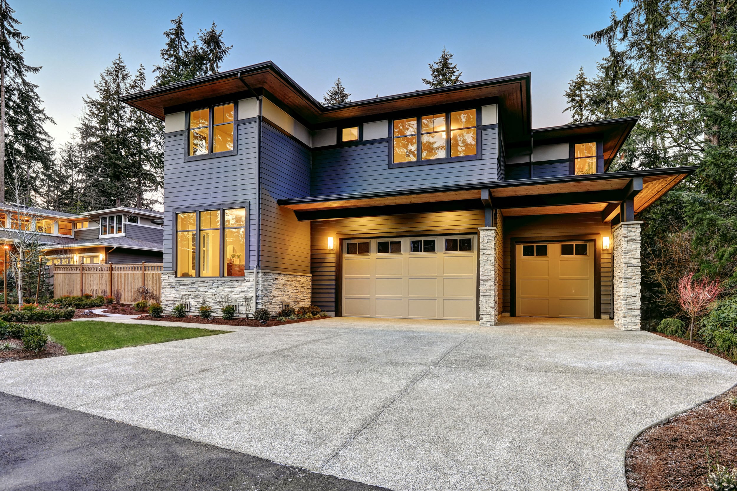 Modern two-story custom home with dark horizontal siding, stone accents, and two-car garage, surrounded by trees and illuminated warmly in early evening.