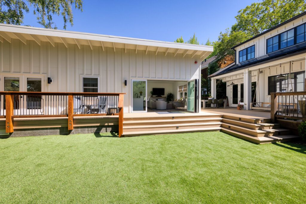 Modern white home with large deck, open glass doors, and artificial grass lawn, adjacent to a second matching structure.