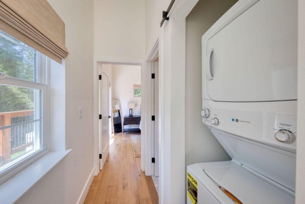 Hallway with window and stacked washer-dryer unit, leading to a bedroom with natural light and hardwood floors.