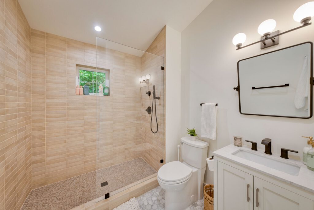 Modern bathroom with a walk-in glass shower, light tan tile walls, white vanity, and black fixtures.