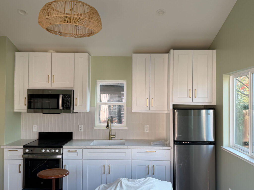 Compact kitchen with sage green walls, white cabinetry with gold handles, a small window over the sink, stainless steel appliances, and a wicker light fixture.