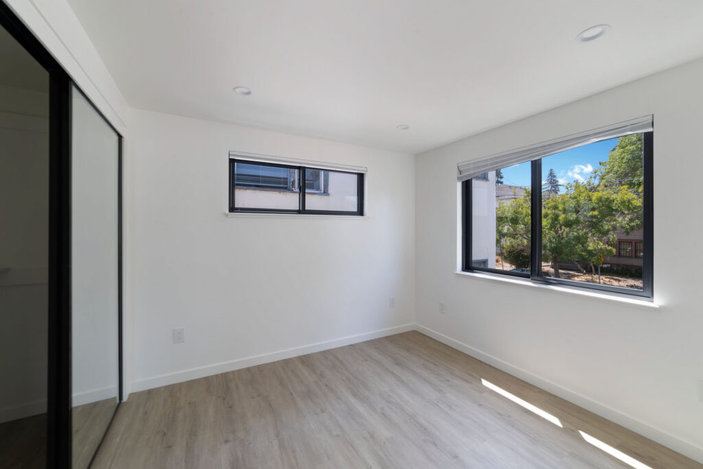 Bright bedroom with large windows, light wood flooring, and mirrored closet doors.