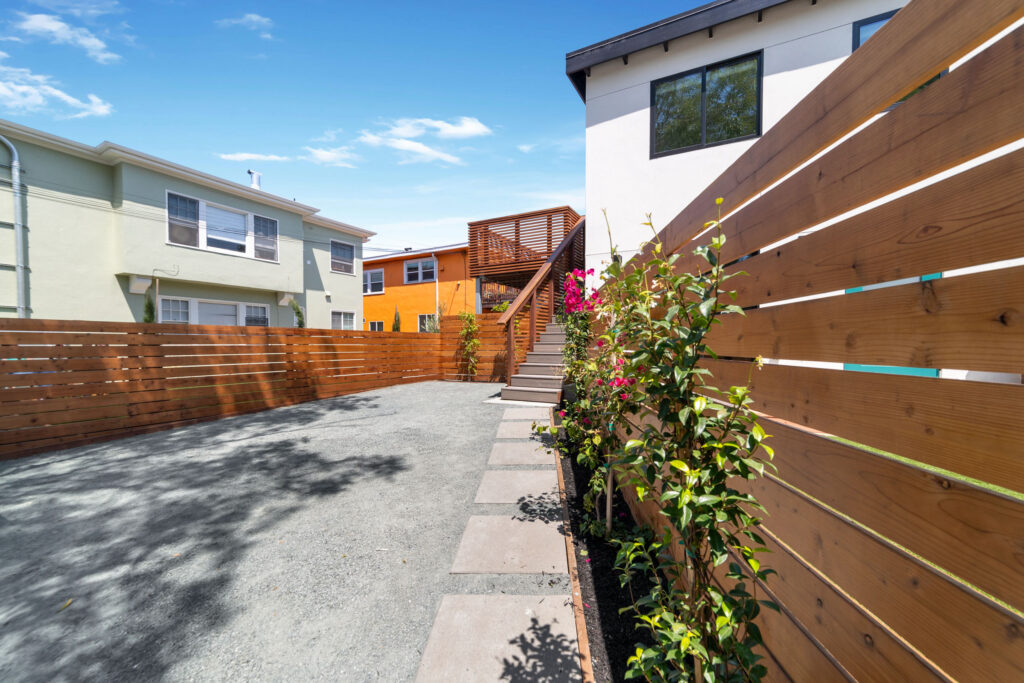Outdoor walkway with paver stones, bordered by a wood fence and flowering plants, leading to a modern staircase and balcony.