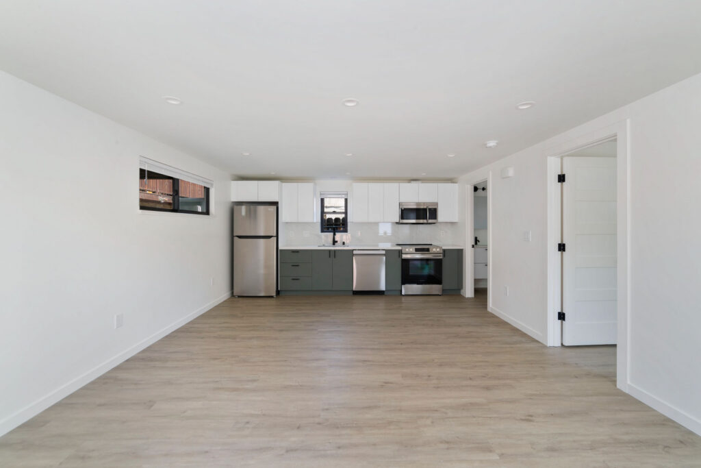 Open-concept kitchen and living space with stainless steel appliances, wood flooring, and minimal white cabinetry.