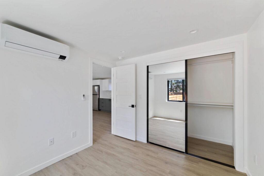 Minimalist bedroom with mirrored sliding closet doors, a wall-mounted air conditioning unit, light wood flooring, and a view into a modern kitchen.