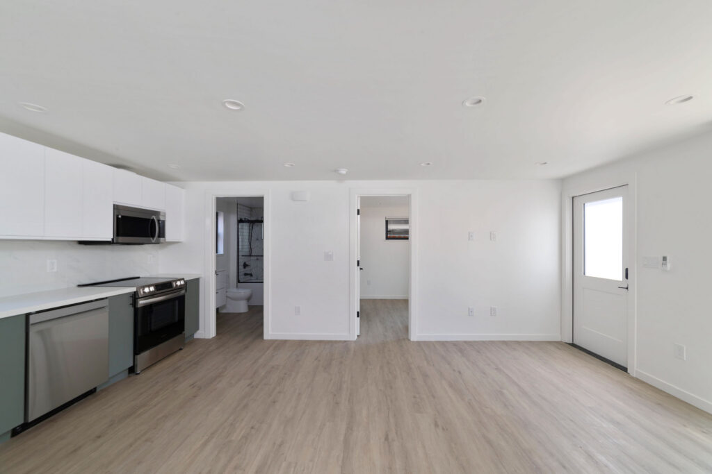 Bright interior view showing a modern kitchen, light wood floors, access to a bathroom with black fixtures, and entry to a separate room through open doors.