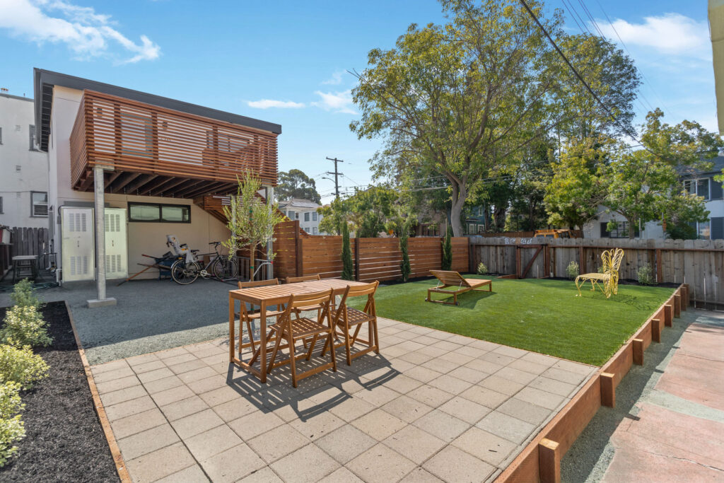 Backyard with patio dining area, artificial turf, loungers, butterfly chair, and a two-story building with a wooden balcony and bike storage underneath.