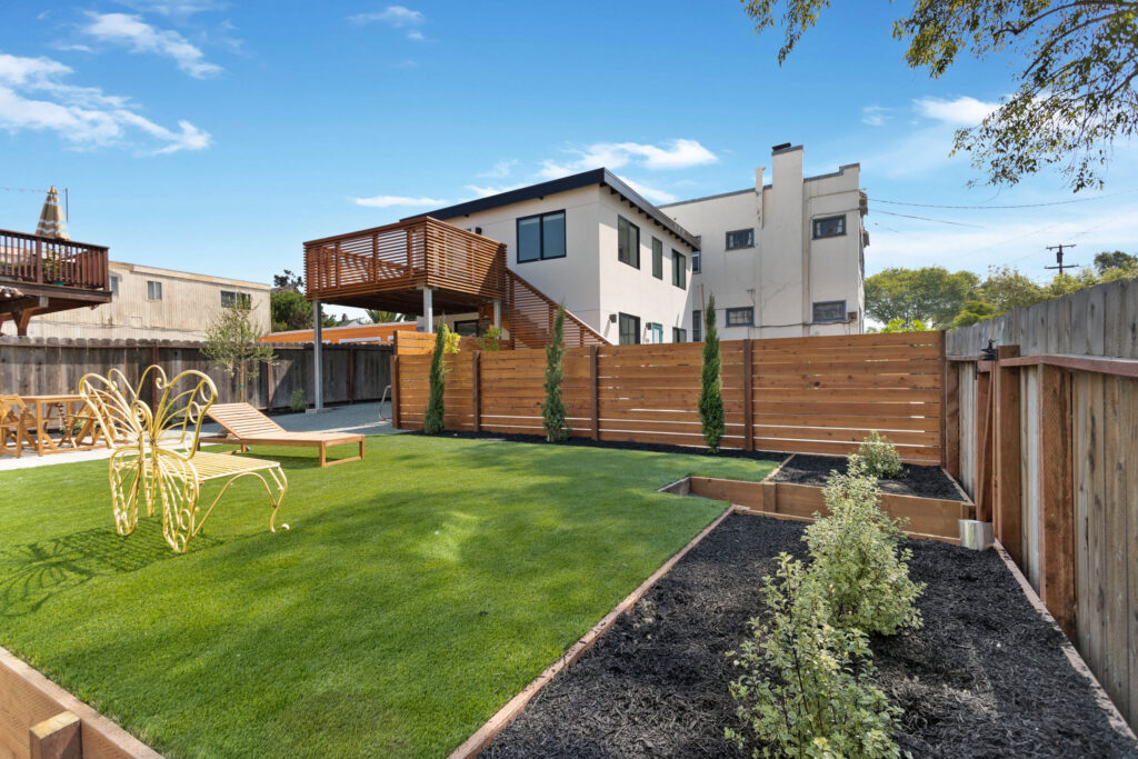 Backyard with green lawn, butterfly-shaped chair, wood loungers, garden beds, and a modern duplex with raised wooden deck and staircase.