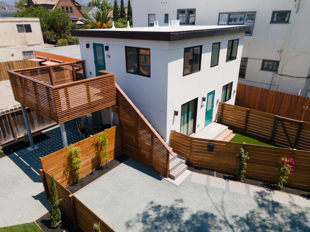 Modern two-story white building with teal doors, wood slat balcony, privacy fencing, landscaped yard, and gravel walkways.