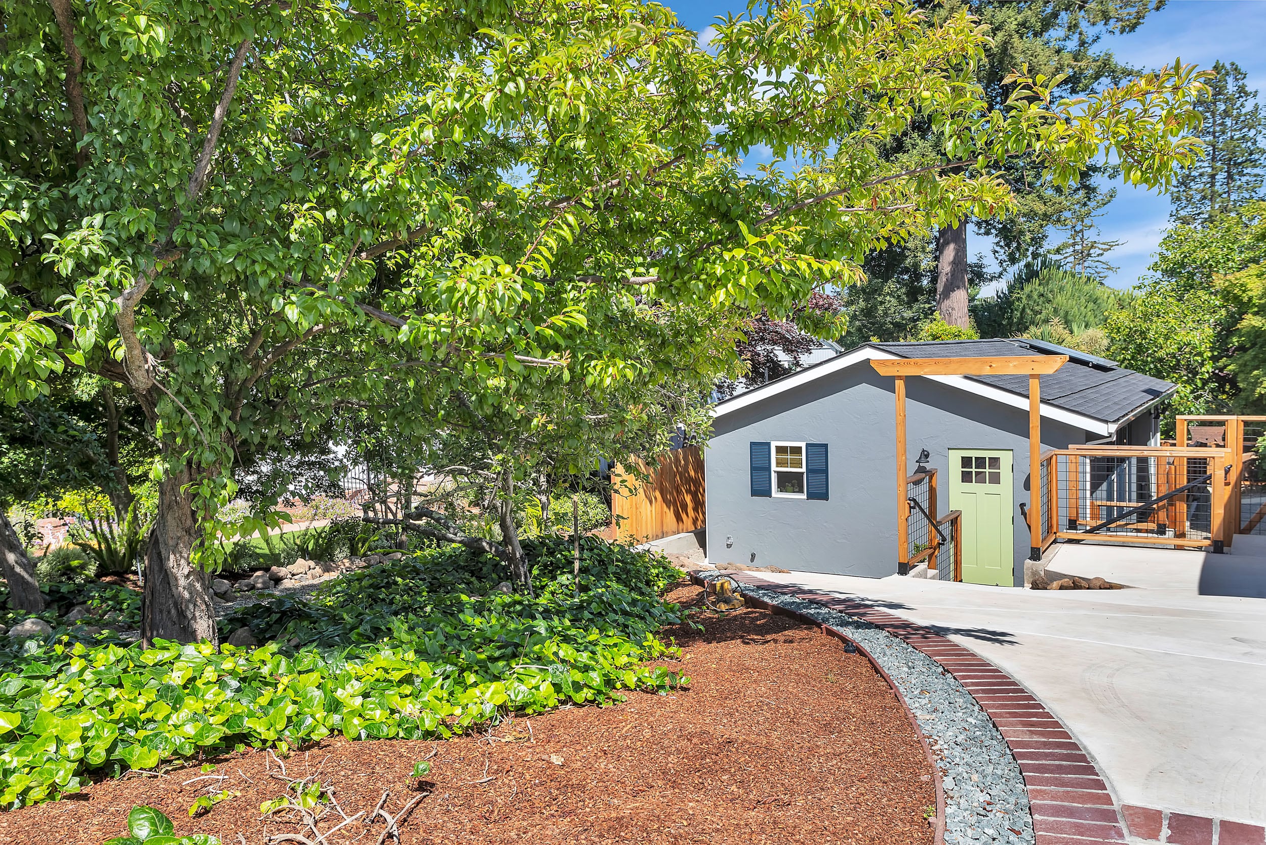 Gray ADU with green door nestled under mature trees, surrounded by lush ivy ground cover and bordered by a landscaped gravel and mulch pathway.